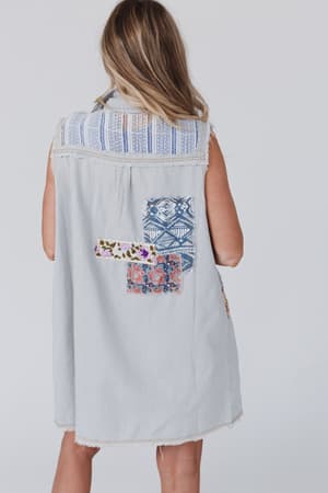 A woman with long, wavy hair models the One Of A Kind Sleeveless Denim Dress Vest - Denim Multi, featuring colorful patchwork and a frayed hem, shown from behind against a plain background.