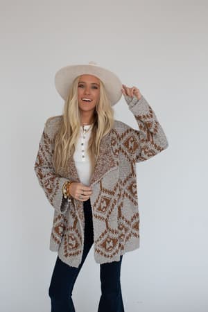 A woman with long blonde hair smiles against a plain white background, wearing the Free to Roam Cardigan in gray over a white shirt, wide-brimmed hat, and dark pants.