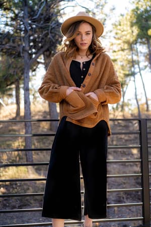 A woman stands outdoors by a metal railing, wearing the Simply Comfort Jumpsuit - Black and a tan hat with a brown jacket. Sunlight and trees fill the background as she looks at the camera with her hands in her jacket.
