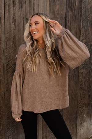 A smiling woman with long, wavy blonde hair wears The Nest Sweet Heart Balloon Sleeve Knit Sweater in mocha and black pants, posing with one hand raised in front of a rustic wooden wall.
