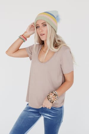 A woman with long blonde hair wears a Scoop Neck Bralette Tee in khaki, a pastel striped beanie, blue velvet pants, and colorful bracelets while posing against a plain white background.