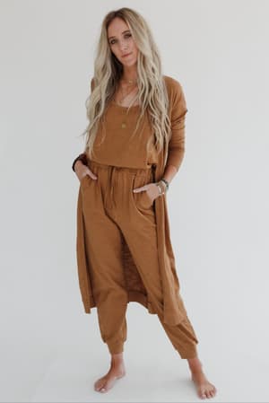 A woman with long, wavy blonde hair stands barefoot against a white background, wearing the Set to Go Textured Jumpsuit And Cardigan Set in camel, styled with layered necklaces and her hands in her pockets.