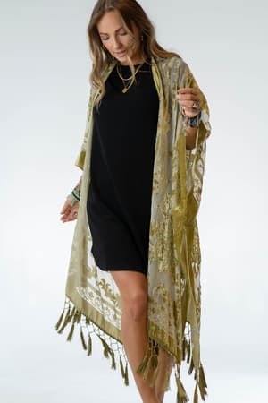 A woman wears a short black dress with the Crushin' On You Velvet Kimono in Olive, featuring sheer fabric, floral patterns, and tassel details. She smiles while looking down, wrists adorned with bracelets, set against a light background.