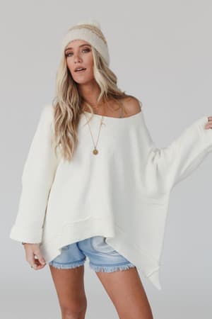 A woman wears the Dana Oversized Diamond Texture Sweater in ivory with a relaxed expression, one shoulder exposed, paired with light blue denim shorts and a cream beanie against a plain background.