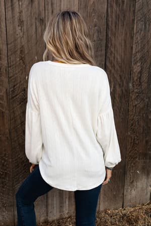 A woman with wavy blonde hair stands facing a wooden fence, wearing The Nest Sweet Heart Balloon Sleeve Knit Sweater in Cream and dark jeans. Her back is to the camera, and the ground is covered with dry grass.