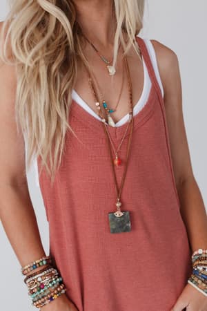 A woman wears a sleeveless rose top over a white tank and the Giselle Stone and Shell Boho Necklace in turquoise, layered with beaded bracelets. Her wavy blonde hair falls loosely over her shoulder.