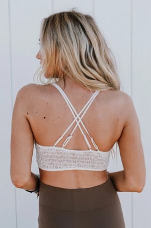 A woman with long blonde hair stands facing away, wearing the Dainty Daisy Crochet Bralette in White and high-waisted brown leggings, set against a light-paneled wall.