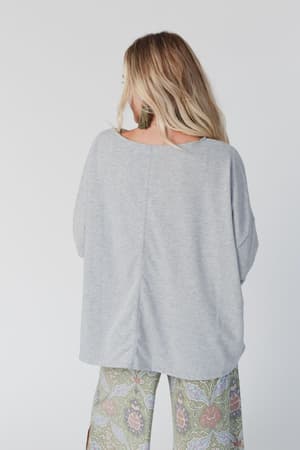 A woman with long, wavy blond hair stands facing away, wearing The Nest Cozy Cool Oversized Tee in Heather Gray and patterned green side-slit pants against a plain light background.