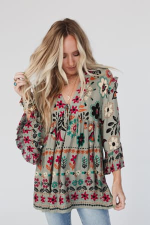 A woman with long, wavy blonde hair models the Leona Floral Print Tunic in Sage, featuring a relaxed fit and soft v-neckline. She pairs it with blue jeans and poses against a plain white background, looking down and slightly to the side.