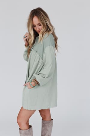 A woman with long, wavy blonde hair wears the oversized Sunday Kind Of Love Dress in sage and gray knee-high boots. She looks down with one hand in her pocket, accessorized with multiple bracelets, against a plain white background.
