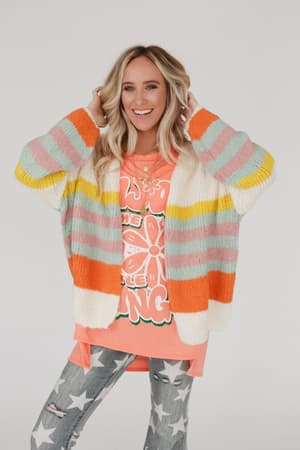 A smiling woman with long blonde hair wears the Retro Babe Striped Cardigan - Cream over an orange graphic tee and distressed gray jeans with white stars, standing against a plain white background.