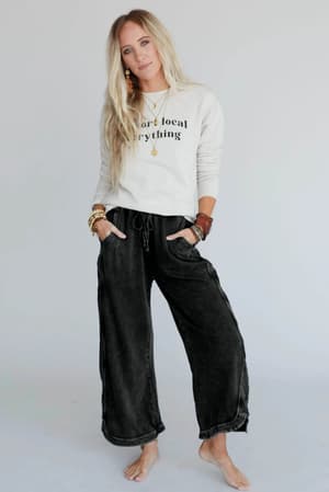 A barefoot woman with long blonde hair stands against a plain background, wearing a white sweatshirt that reads More local everything and So Comfy Wide Leg Pant Cropped Length - Charcoal, accessorized with layered necklaces and bracelets.