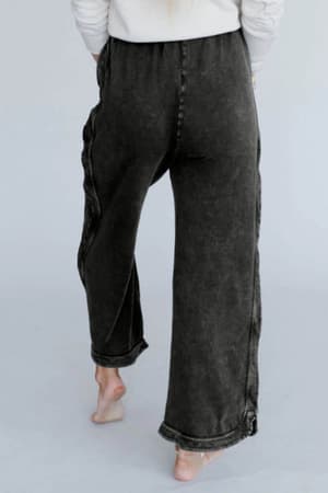 A person stands barefoot on a light surface, shown from the back, wearing high rise elastic waist So Comfy Wide Leg Pant Cropped Length in charcoal with a wide leg silhouette and a long-sleeve white top.