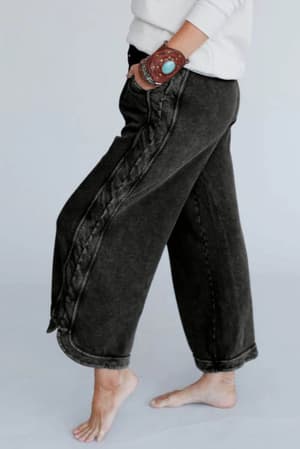 A person stands barefoot, hand in pocket, wearing the So Comfy Wide Leg Pant Cropped Length in Charcoal with a relaxed wide-leg fit and decorative side seams. A turquoise bracelet and a plain backdrop enhance the outfit's laid-back vibe.