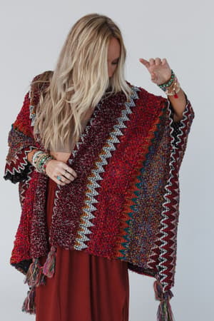 A woman with long blonde hair models the Desert Dreams Crochet Ruana - Maroon, styled over a maroon dress. She looks down with her arm raised, creating a boho vibe against a simple light background.
