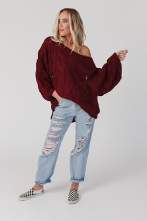 A woman with blonde hair stands against a white background, wearing the Golden Hour Bubble Sleeve Sweater Dress in burgundy, styled off-the-shoulder with ripped light blue jeans and checkered slip-on shoes.