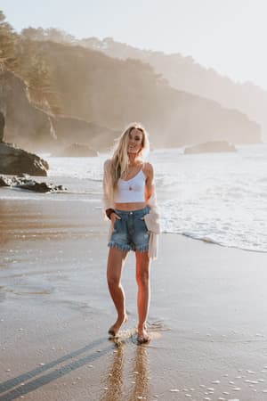 Barefoot on a sandy beach with waves and cliffs behind her, a woman with long blonde hair wears a white crop top, denim shorts, and the Brenden Knit Netted Cardigan in Ivory.