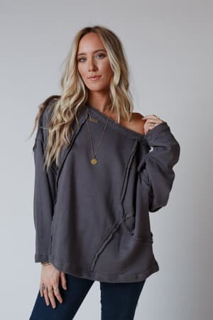 A woman with long blonde hair models the Bailey Oversized Raw Edge Top in charcoal and dark jeans against a plain backdrop. She has minimal jewelry, a relaxed expression, and exudes a subtle boho style.
