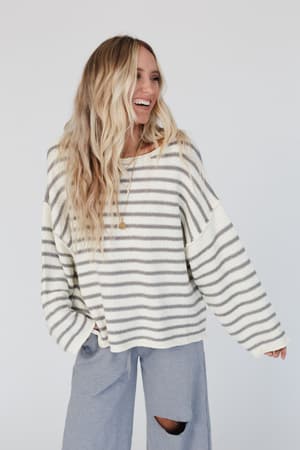 A woman with long, wavy blonde hair smiles in the All My Stripes Oversized Sweater - Ivory Gray and light gray ripped-knee pants, standing against a plain white background.