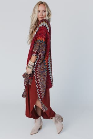A woman with long blonde hair wears the Desert Dreams Crochet Ruana in Maroon—a colorful, textured shawl with zigzag patterns—over a rust dress and beige fringe ankle boots, standing against a plain white background.