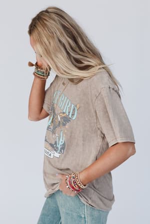 A woman with long blonde hair stands sideways, looking down and touching her hair while wearing an oversized Fly Free Graphic Tee - Mocha, blue jeans, and several colorful boho-chic beaded bracelets on her wrist.