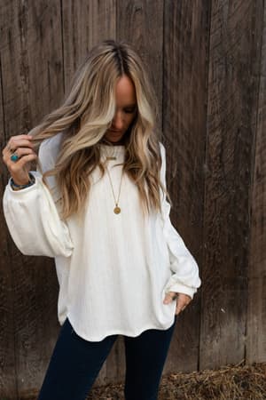 A woman with long, wavy blonde hair wears The Nest Sweet Heart Balloon Sleeve Knit Sweater in cream and dark jeans, standing before a weathered wooden wall as she looks down and touches her hair.