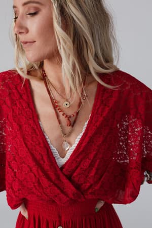 A woman with loose blonde hair wears a red lace top over a white bralette, styled with the Carnelian Multi Layered Necklace - Rust and beaded jewelry, gazing to the side against a plain background.