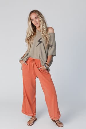 A woman with long blonde hair stands confidently against a plain light background, wearing a beige off-the-shoulder top, the So Comfy Wide Leg Pant Cropped Length in Faded Rust, multiple bracelets, and sandals.