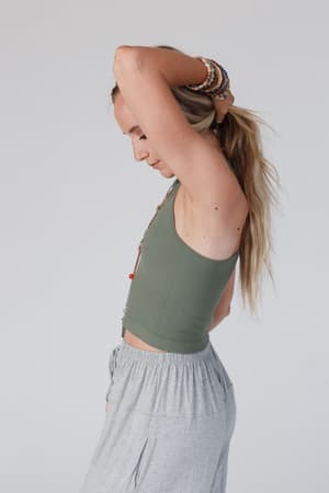 A woman with long blonde hair tied back wears the High Neck Racerback Brami in Olive, paired with gray pants and beaded bracelets. She stands in profile, one arm raised to her head, against a plain light-colored background.
