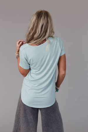 A woman with long, wavy blonde hair stands with her back to the camera, wearing a Scoop Neck Bralette Tee in Washed Seafoam and loose gray pants—perfect for layering—against a plain gray background.