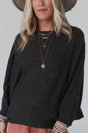 A woman with long blonde hair stands against a plain light background, wearing The Nest Fly With Me Batwing Sleeve Knit Sweater in charcoal, a wide-brimmed hat, layered gold necklaces, and several beaded bracelets.