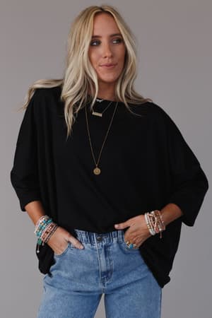 A woman with long blonde hair wears The Nest Cozy Cool Oversized Tee in black, paired with light blue jeans, layered necklaces, and colorful beaded bracelets, standing against a plain gray background with her hands in her pockets.