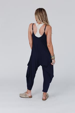 A woman with long blonde hair faces away, wearing The Perfect Harem Jumpsuit - Navy over a white tank top, paired with beige slip-on shoes and bracelets on her left wrist against a plain light background.