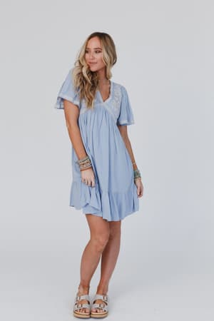 A woman wears the Magdaline Mini Dress - Blue, featuring light blue fabric with white embroidery. She pairs it with sandals and multiple bracelets, her long wavy hair enhancing her effortless look against a plain background.