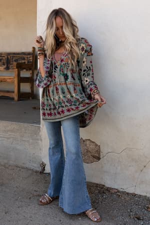A woman with long wavy hair wears the Leona Floral Print Tunic in sage, paired with light blue flared jeans and studded sandals. She stands outdoors against a textured wall, holding her blouse's side and looking down.