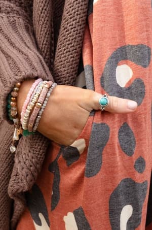 A person in a brown knit cardigan and an orange abstract-pattern dress stands with their hand in their pocket, displaying boho jewelry—several beaded bracelets and the Kapalua Bay Adjustable Turquoise Ring in silver.