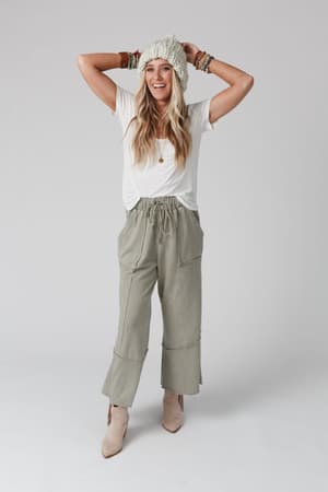 Woman smiling with hands on her head, wearing the Feeling Good Wide Leg Pant in Faded Sage, a white T-shirt, knit beanie, ankle boots, and several bracelets for a relaxed boho style against a plain white background.