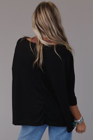 A person with long, wavy blonde hair is seen from behind wearing The Nest Cozy Cool Oversized Tee in black, light blue jeans, and colorful beaded bracelets. The background is plain and neutral.