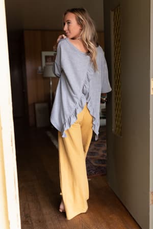 A woman stands barefoot indoors, facing away, wearing The Nest Must Have High Low Tee in heather gray with ruffle accents and flowy yellow pants. The softly lit room has wooden floors and cozy, warm-toned decor in the background.