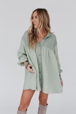 A woman with long, wavy blonde hair wears the Sunday Kind Of Love Dress in sage and knee-high taupe boots. She smiles and looks to the side against a plain white background.