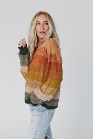 A woman with long blonde hair wears the Going Places Gradient Stripe Sweater - Mustard in orange, yellow, green, and beige stripes. She stands sideways with one hand in her pocket, looking over her shoulder, paired with light blue ripped jeans.