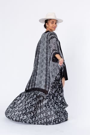 A woman wearing the Paisley Tapestry Free Flow Duster Kimono in black and a wide-brimmed hat poses against a white background, glancing over her shoulder with a subtle smile.