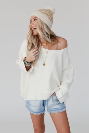 A woman with long blonde hair, wearing a knit beanie, denim shorts, layered necklaces, bracelets, and the Dana Oversized Diamond Texture Sweater in Ivory off the shoulder, smiles and looks to the side against a plain light background.