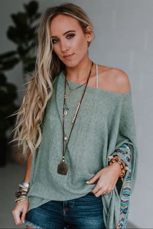 A woman with long, wavy blonde hair wears the Fallon Embroidered Sleeve Poncho in sage with colorful patterned sleeves, layered necklaces, bracelets, and denim shorts. She stands indoors against a blurred background with a plant.