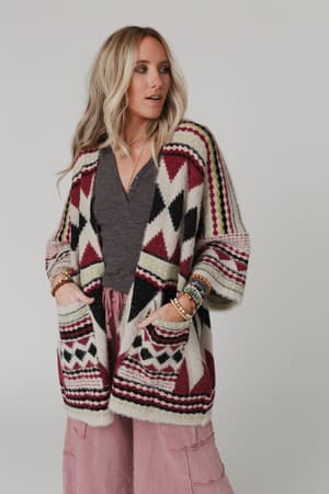 A woman with long, wavy blonde hair wears the Keep It Up Oversized Cardigan - Berry in red, black, and cream over a dark gray top and pink wide-leg pants, accessorized with multiple bracelets and her hands in her pockets.
