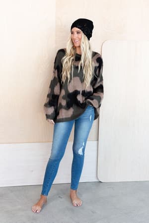 A woman with long, wavy blond hair wears the Wild Wanderer Sweater - Camo, a black beanie, and ripped skinny jeans. She stands barefoot on a concrete floor against a light wood background, smiling and looking to the side.