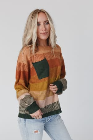 A woman with long blonde hair wears the Going Places Gradient Stripe Sweater in mustard, featuring earth-tone stripes and a green pocket, paired with light blue ripped jeans against a plain light background.