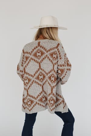 A person wearing the Free to Roam Cardigan - Gray, featuring an Aztec-inspired print, a white wide-brimmed hat, and dark jeans stands against a plain white background facing away from the camera.