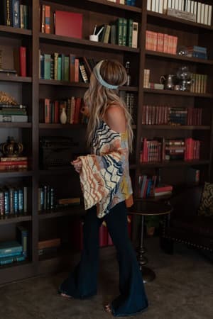 A woman with long hair, wearing the Rainbow Shores Knit Sweater - Multi with flare sleeves, flared jeans, and a headband, stands by a bookshelf looking to the side as she reaches for a book.