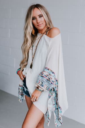 A woman with long blonde hair wears the Fallon Embroidered Sleeve Poncho in ivory, paired with denim shorts, layered necklaces, and colorful bracelets, standing against a white brick wall.